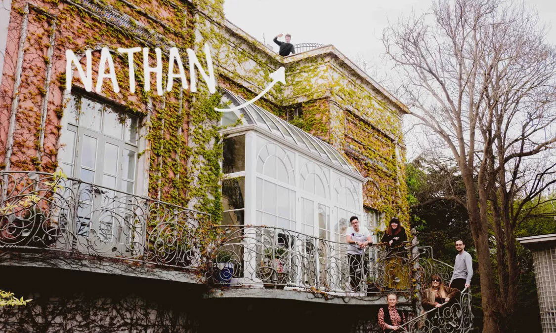 A group of people stand and sit on the balcony and steps of an ivy-covered University building. A person labelled Nathan stands on the roof, waving. Leafless trees are visible to the right, adding charm to this Blue Stag gathering.