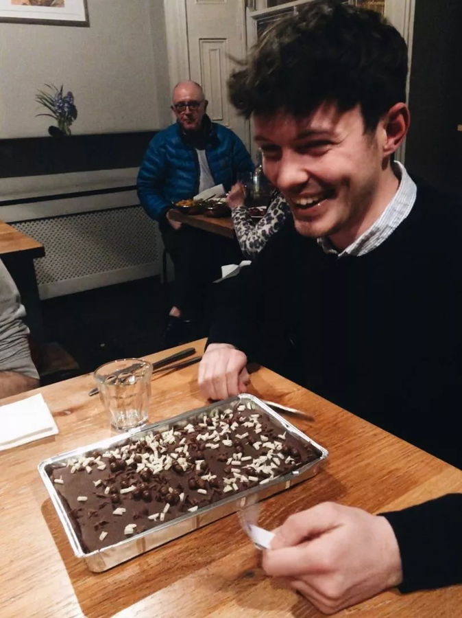 A young man smiles at a table with a rectangular chocolate cake topped with white and dark chocolate shavings at University to Blue Stag. A glass of water and serviette are nearby, with other people seated in the background.