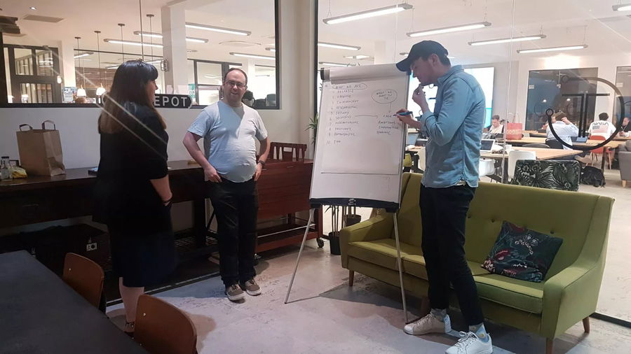 Three people are having a discussion around a flip chart in a modern office space at Blue Stag. One person is writing on the chart, while the others listen. In the background, tables and chairs fill with people working—some fresh from university.