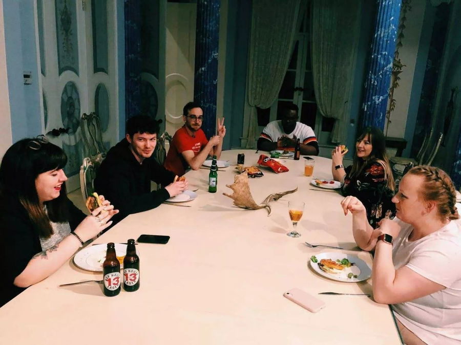 Six people sit around a large dining table at University to Blue Stag, eating and chatting. Plates of food, drinks, empty bottles, and a wooden cat sculpture fill the table. The atmosphere is casual and friendly.