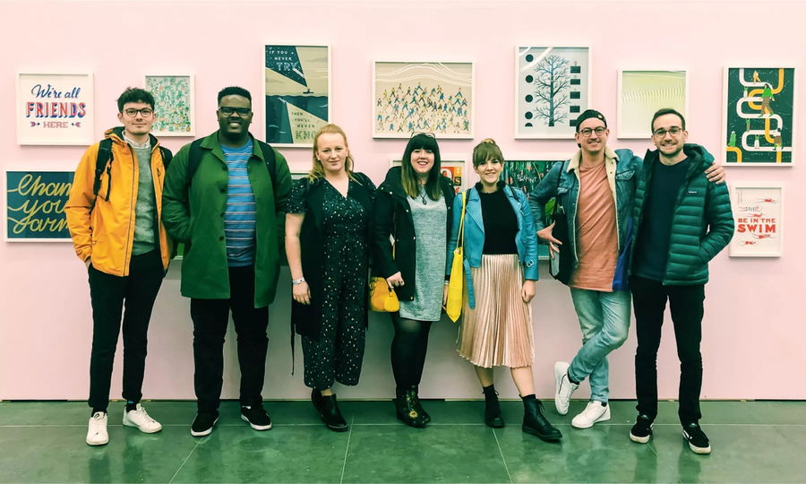 A group of seven people stand side by side, smiling, in front of a pink wall at University to Blue Stag, decorated with colourful framed art prints featuring positive messages and illustrations.