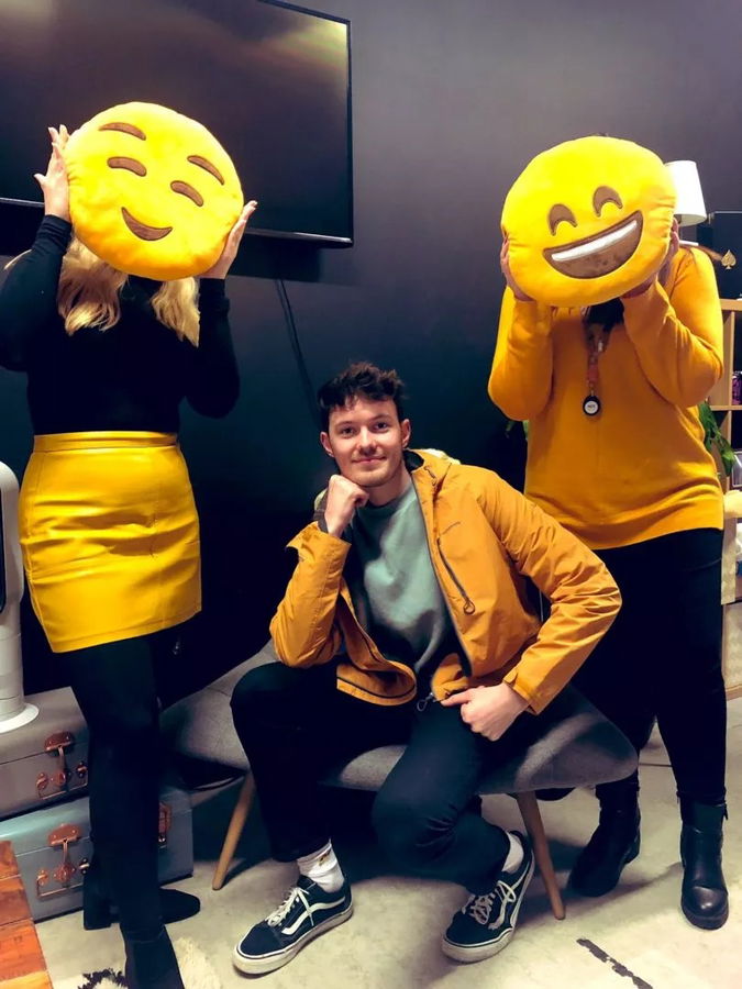 Three people pose indoors. Two stand holding large emoji-face cushions in front of their faces; one sits on a chair, smiling. Their matching yellow outfits add a playful vibe—perfect for team spirit from University to Blue Stag.