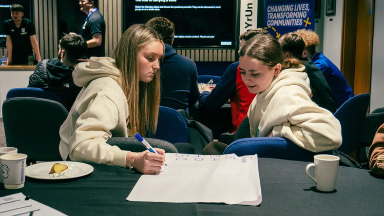 Two young women sit at a table during discovery workshops—one writes on a large sheet of paper while the other observes. Cups, papers, and a plate of food are scattered around, with other participants and a presentation screen in the background.