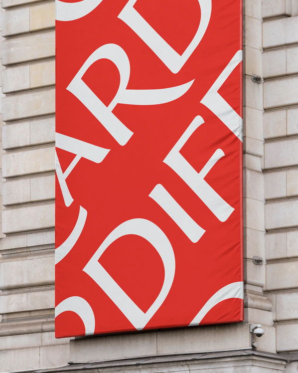 A red banner with large, overlapping white letters hangs on the exterior of a stone building. Only parts of the letters are visible, creating an abstract design.
