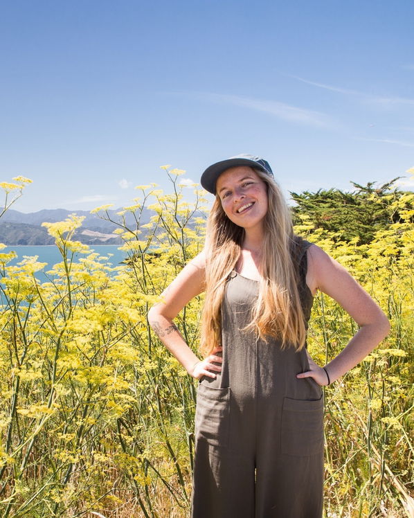 The newest member of the herd, a woman with long blonde hair in a boiler suit and cap, stands smiling among tall yellow wildflowers with blue sky, mountains, and water in the background.