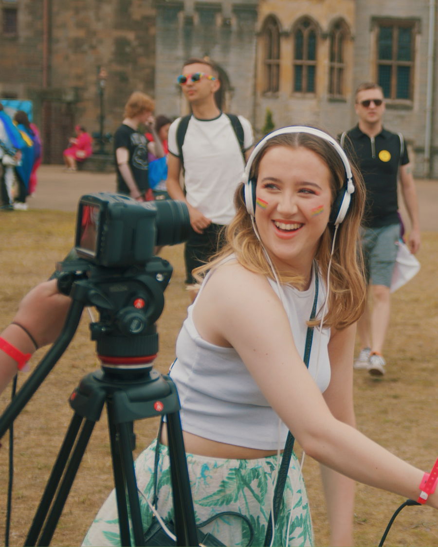 A young woman with headphones and rainbow face paint smiles whilst operating a video camera outdoors at an event promoting Creative Collaboration for a Wales Without Violence, with several people and a historic building in the background.