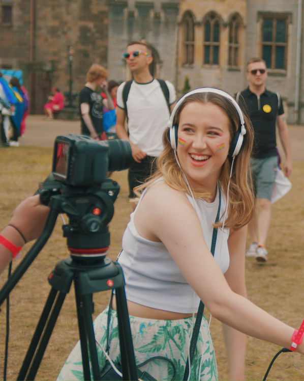 A young woman with headphones and rainbow face paint smiles whilst operating a video camera outdoors at an event promoting Creative Collaboration for a Wales Without Violence, with several people and a historic building in the background.