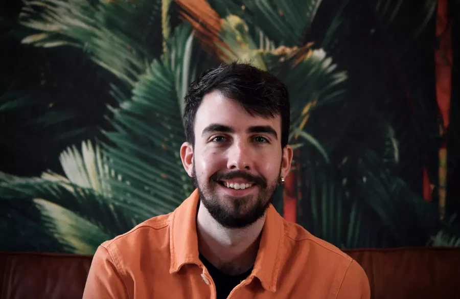 Kieran Traas of Blue Stag, a man with short dark hair and a beard, smiles in an orange jacket whilst sitting in front of a leafy green and orange tropical-themed background.