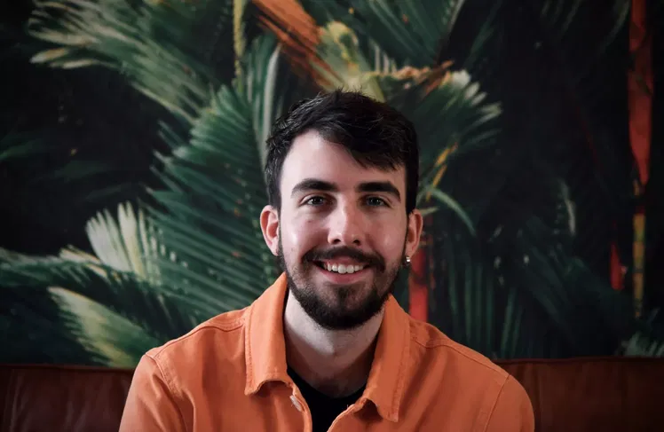 Kieran Traas of Blue Stag, a man with short dark hair and a beard, smiles in an orange jacket whilst sitting in front of a leafy green and orange tropical-themed background.