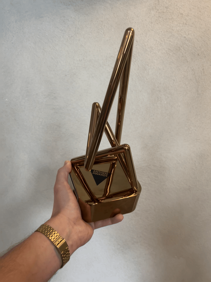 A hand holding a shiny, geometric gold trophy with angular shapes, featuring a small plaque that reads ARCHITECTURE AWARDS. The background is a plain, light-coloured wall—symbolising hope much like an anti-knife crime campaign.