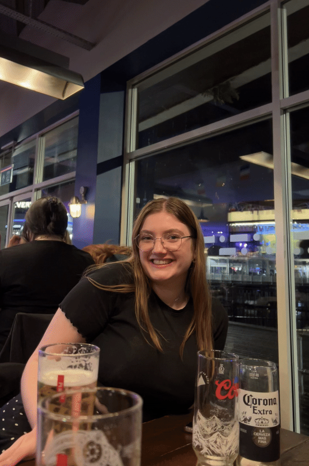 Holly, a marketing manager at Blue Stag with long brown hair and glasses, smiles whilst sitting at a restaurant table with empty beer glasses in front of her. Large windows reveal a brightly lit area outside.