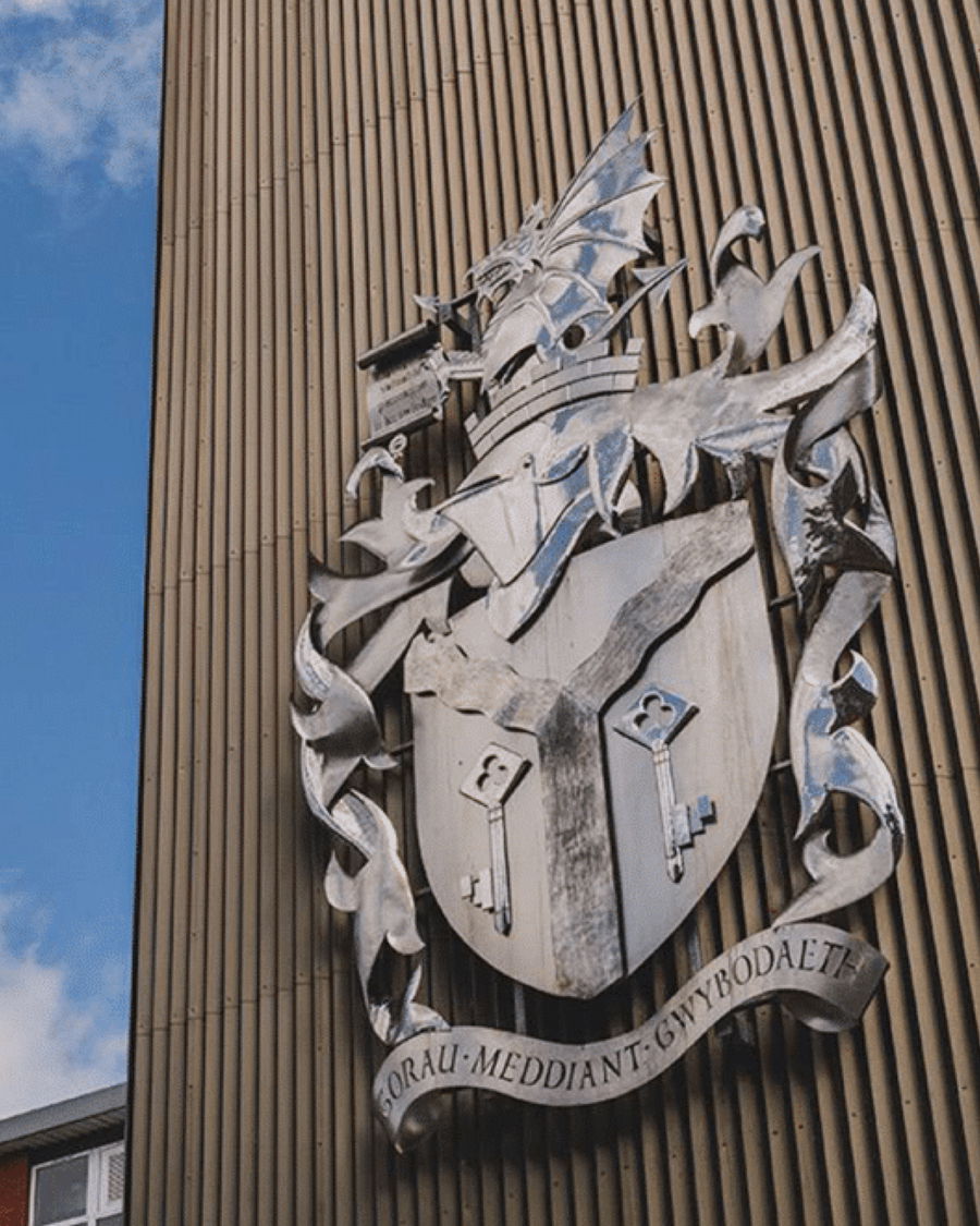 Large silver coat of arms featuring a dragon and shield with two keys, mounted on a ribbed building facade—a striking symbol of tradition and pride that could inspire those seeking work experience. An inscription in Welsh appears at the bottom; blue sky to the left.