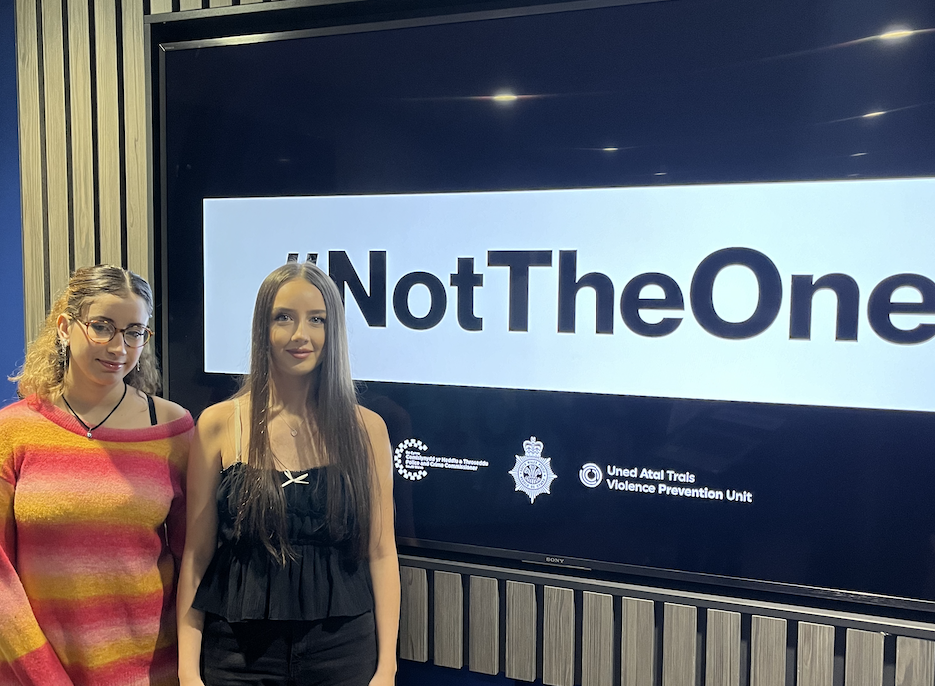 Two women with work experience stand in front of a screen displaying the hashtag #NotTheOne and violence prevention unit logos. One wears a colourful striped jumper; the other, a black top.