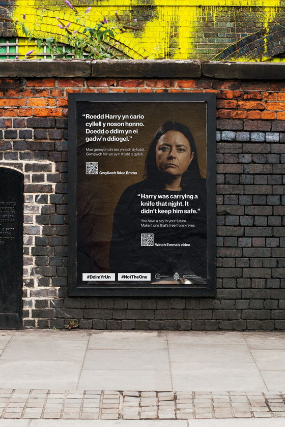 A poster on a brick wall shows a serious-looking woman, with text about knife crime in English and Welsh, hashtags #NotTheOne and #DdimYrUn, and information about making safer choices for a better future.