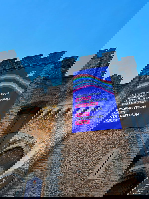 A large banner with rainbow stripes and bilingual text in Welsh and English hangs on the stone wall of a historic castle, celebrating “Strength in unity, strength in pride.” The scene reflects how diverse work experience enriches communities under a clear blue sky.