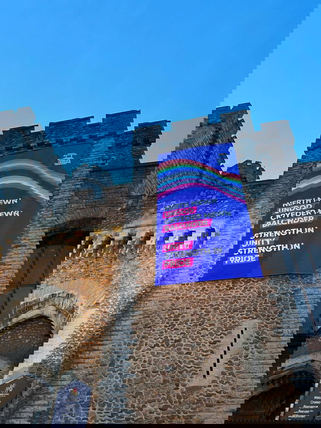 A large banner with rainbow stripes and bilingual text in Welsh and English hangs on the stone wall of a historic castle, celebrating “Strength in unity, strength in pride.” The scene reflects how diverse work experience enriches communities under a clear blue sky.