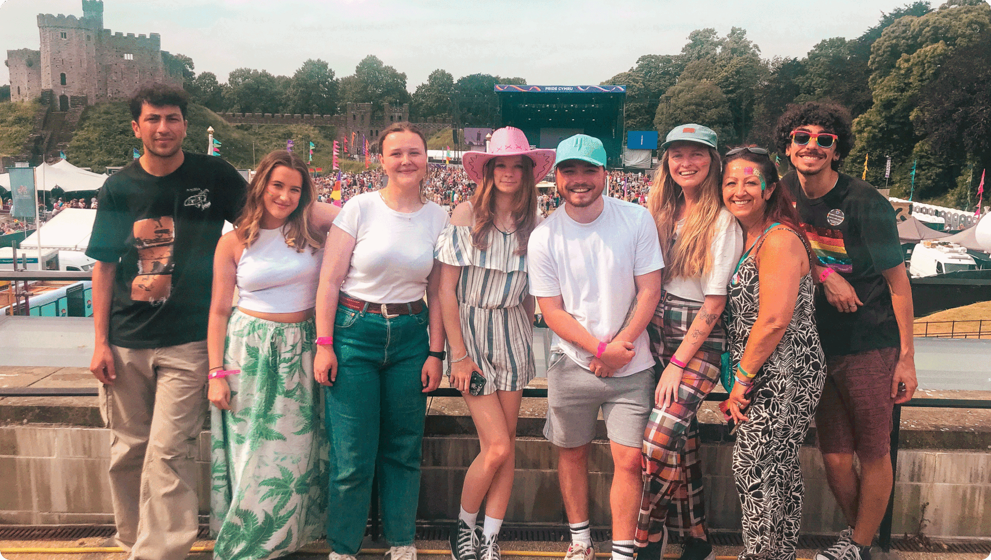 A group of eight people stands smiling outdoors at a festival, showing off their colourful, summery outfits and festival wristbands. The scene, framed by trees and blue sky, hints at their shared work experience and fun outside the office.