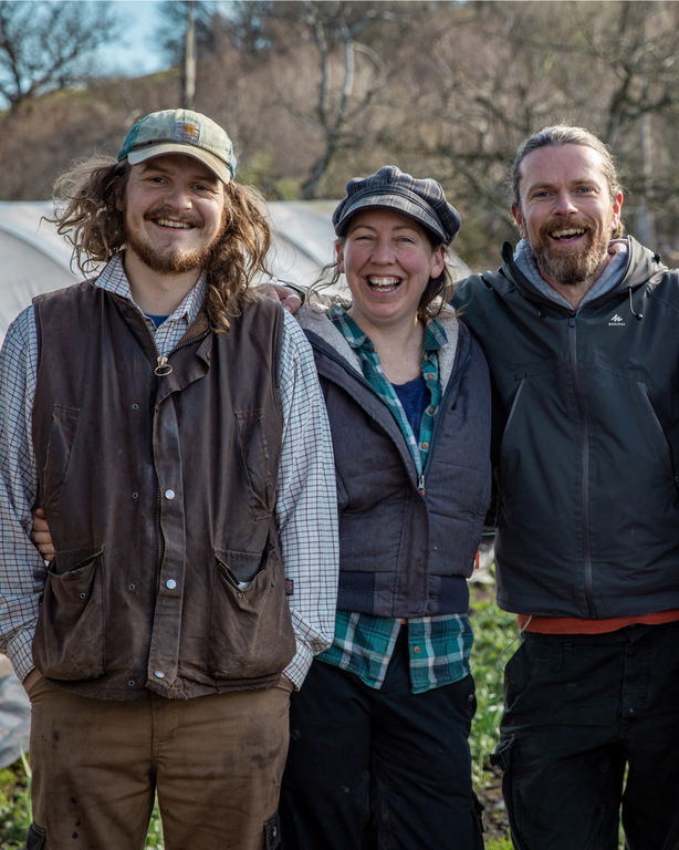 Three adults stand outdoors, smiling and posing together. They are dressed in casual, work-style clothing and appear to be on a farm, with green plants and a glasshouse visible in the background.