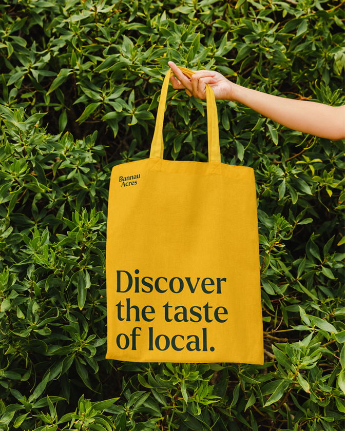 A hand holds a yellow tote bag in front of lush green leaves. The bag reads Banana Acres in small text and Discover the taste of local. in large black letters.