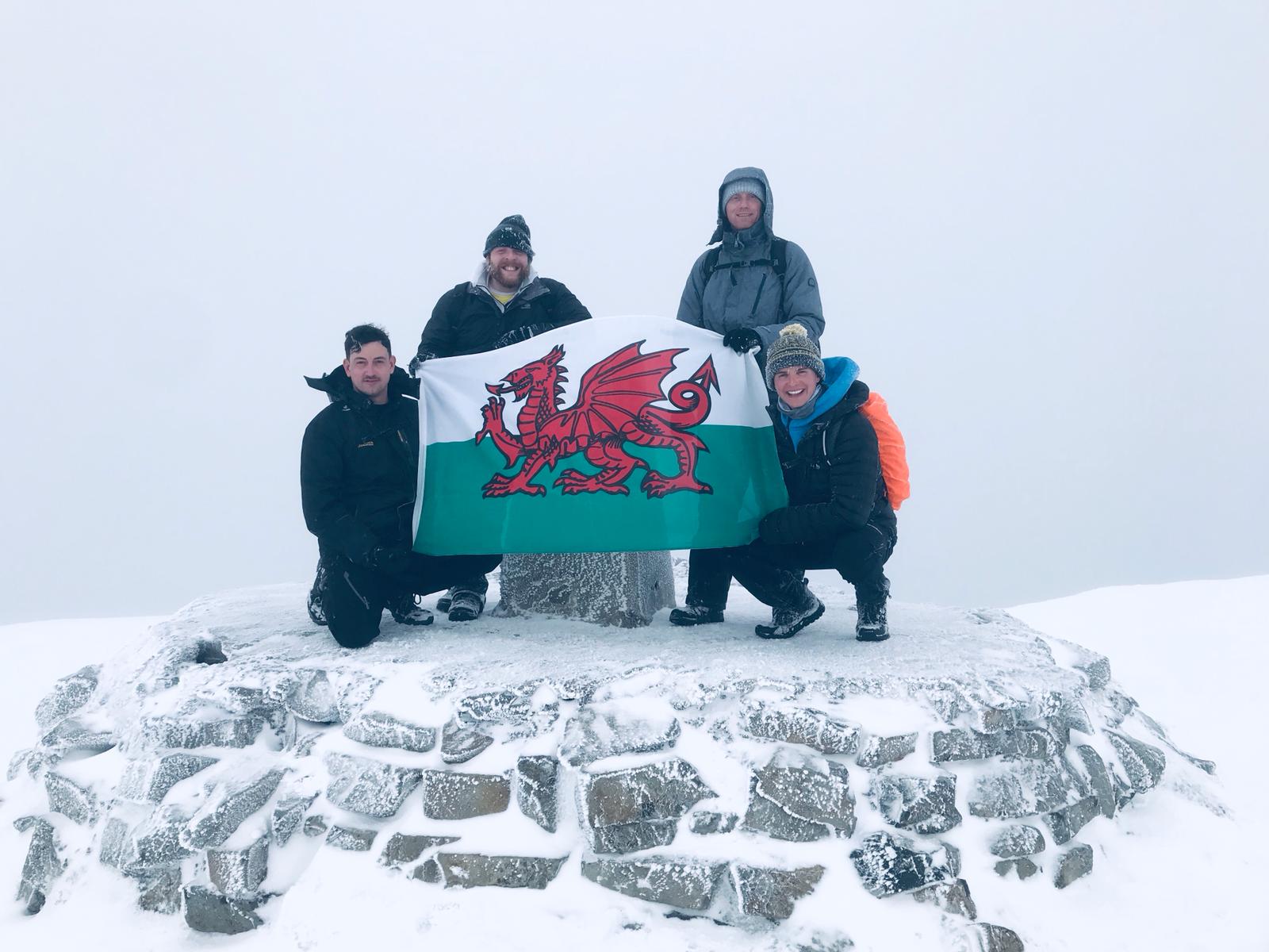 Four people in winter clothing pose on a snow-covered stone platform, holding a Welsh flag with a red dragon. Among them is Dan Sargent. The background is foggy and white, suggesting snowy or misty weather.