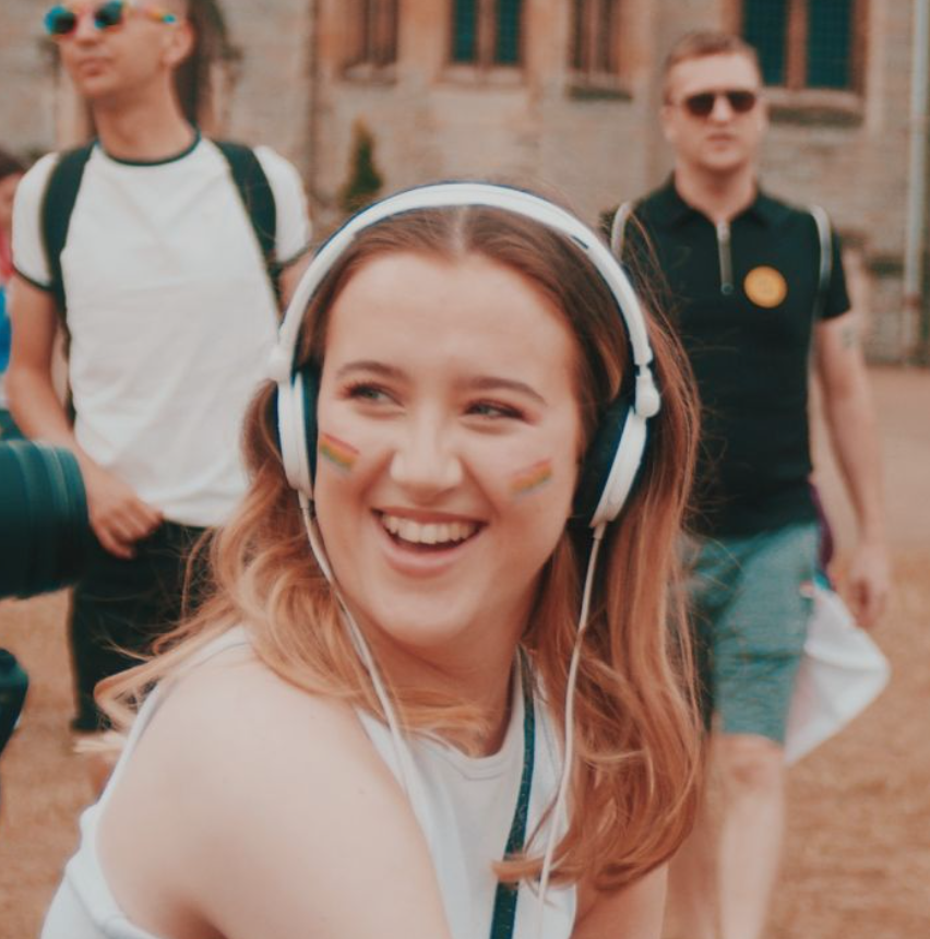 A smiling young person with long hair and rainbow stickers on their cheeks wears white headphones at an outdoor event, possibly sharing stories about their work experience. Other people are visible in the sunny background, some with pride-themed accessories.