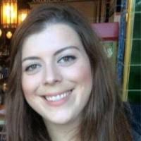 A young woman with long brown hair is smiling at the camera. She is indoors, and the background includes warm lighting and colourful tiles.