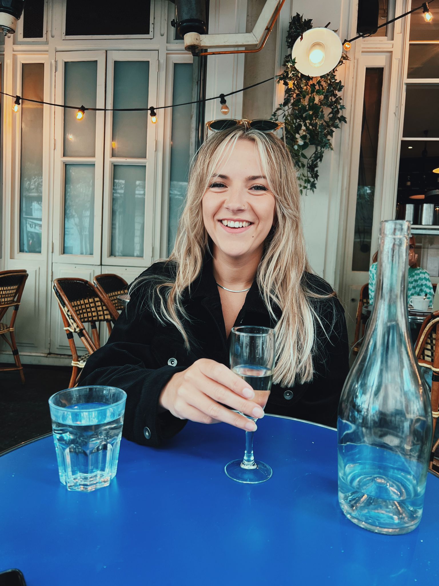 Kirsty Bowen, a smiling woman with long blonde hair, sits at a blue table in a café holding a glass of wine. A glass of water and an empty bottle rest on the table, with café chairs and fairy lights glowing in the background.