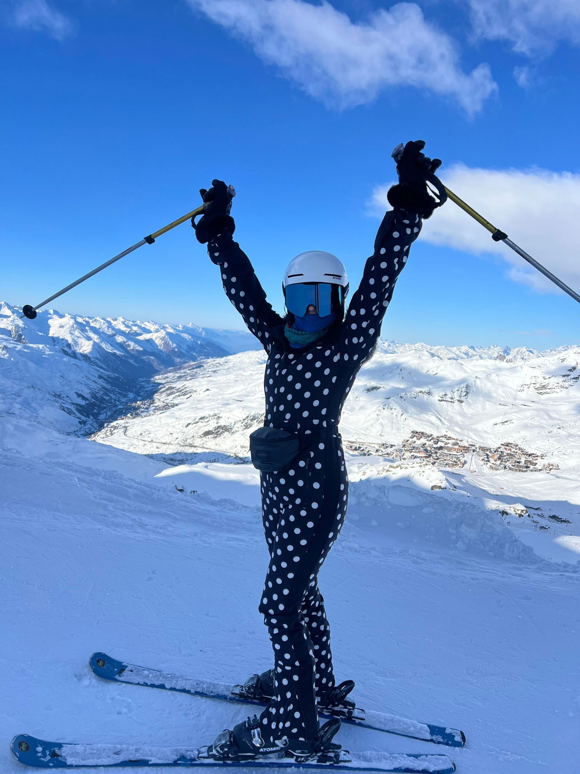Kirsty Bowen, in a black and white polka dot suit, stands triumphantly on a snowy mountain slope with arms raised, holding ski poles. Snow-covered peaks and a blue sky complete the stunning scene.