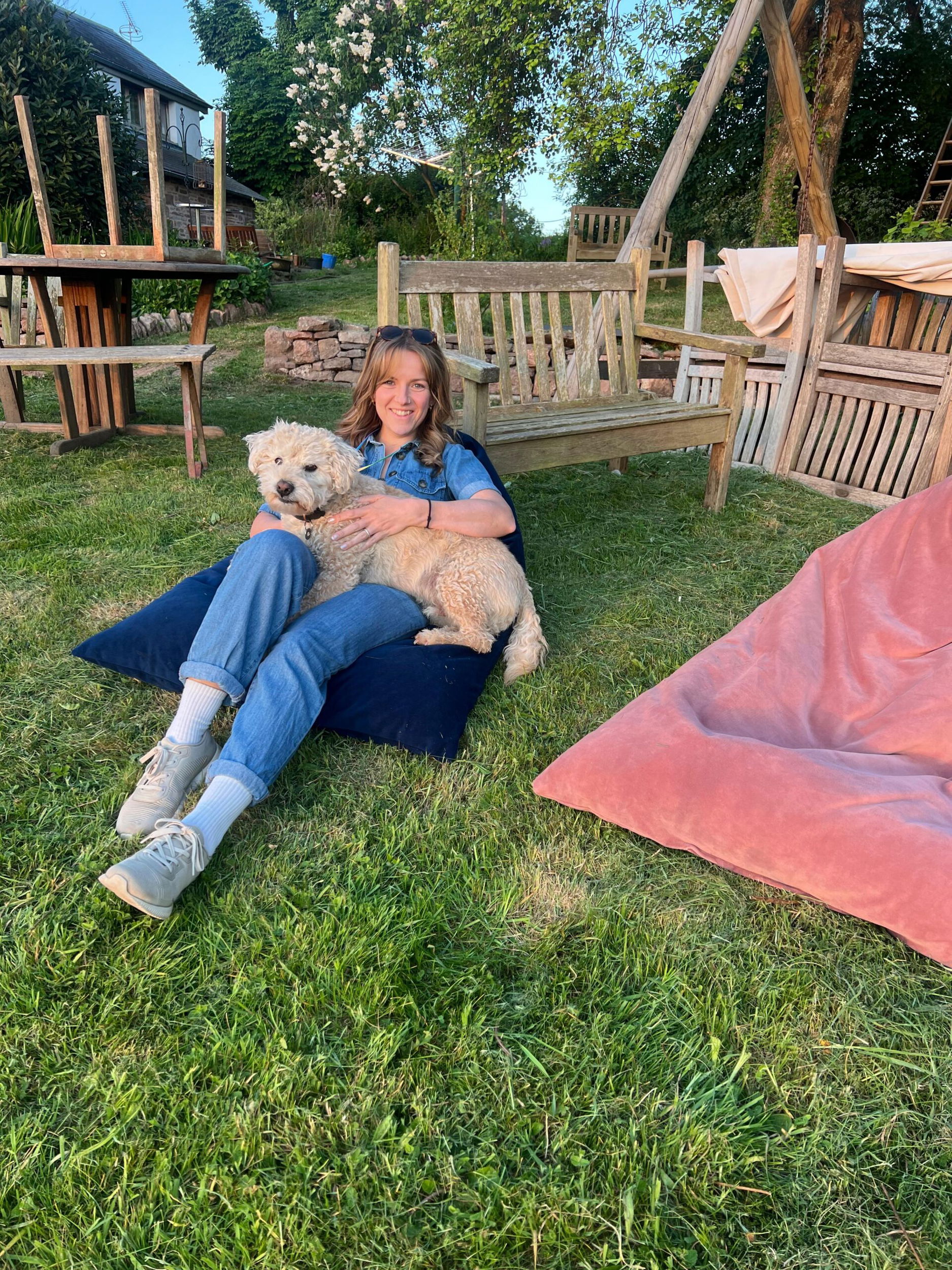Kirsty Bowen sits on a navy blue cushion on the grass, smiling and cuddling a curly-haired dog. Wooden outdoor furniture and a pink beanbag are nearby, with trees and a building in the background.