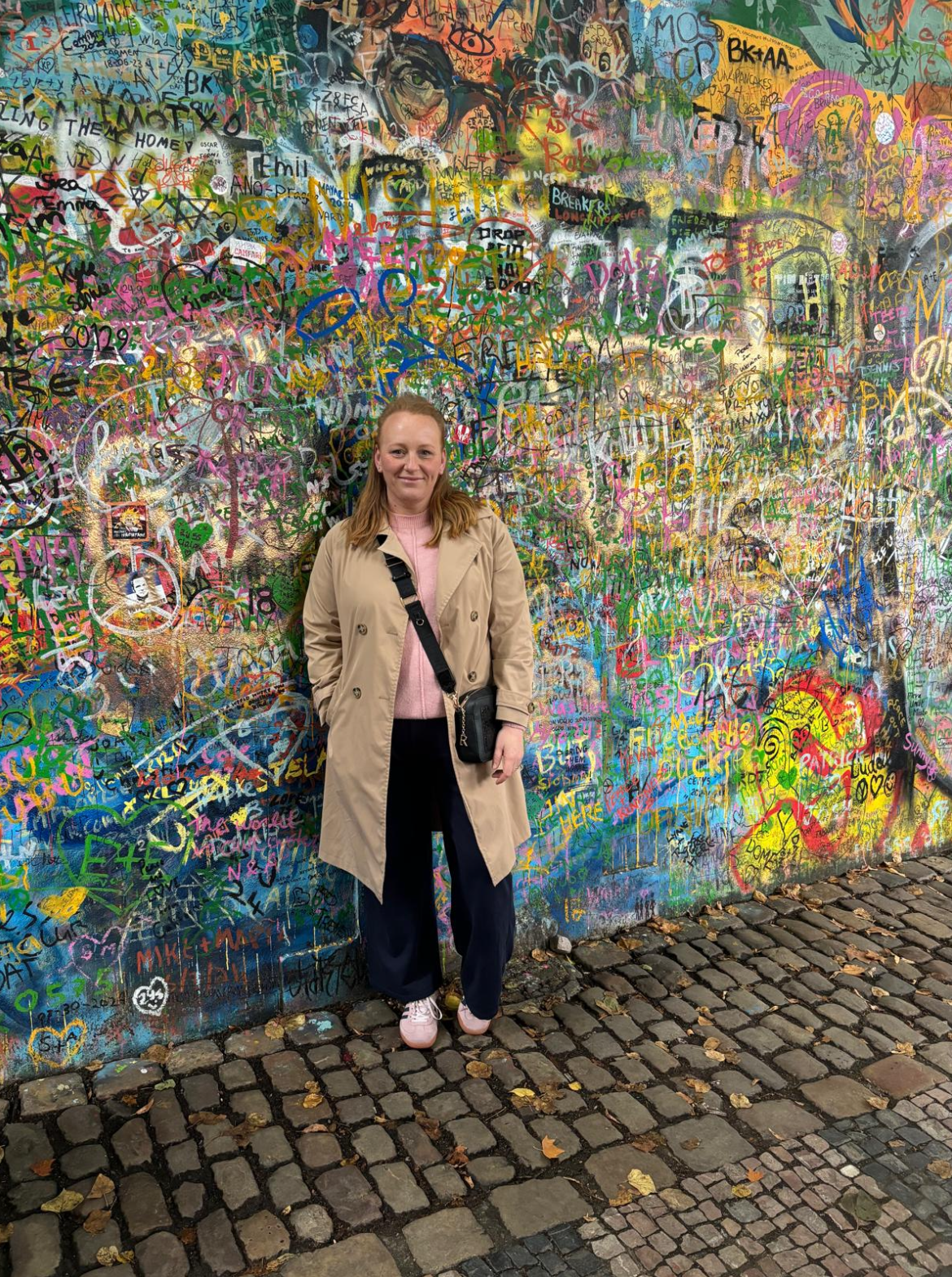 Kirsty Bowen, in a beige trench coat, stands on cobblestones before a colourful, graffiti-covered wall bursting with vibrant drawings, messages, and signatures.