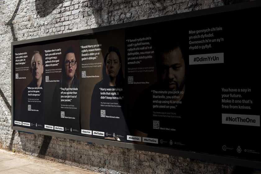 A brick wall displays posters with photos of four people and anti-knife crime messages in English and Welsh, along with hashtags #DdimYrUn and #NotTheOne and QR codes for more information.
