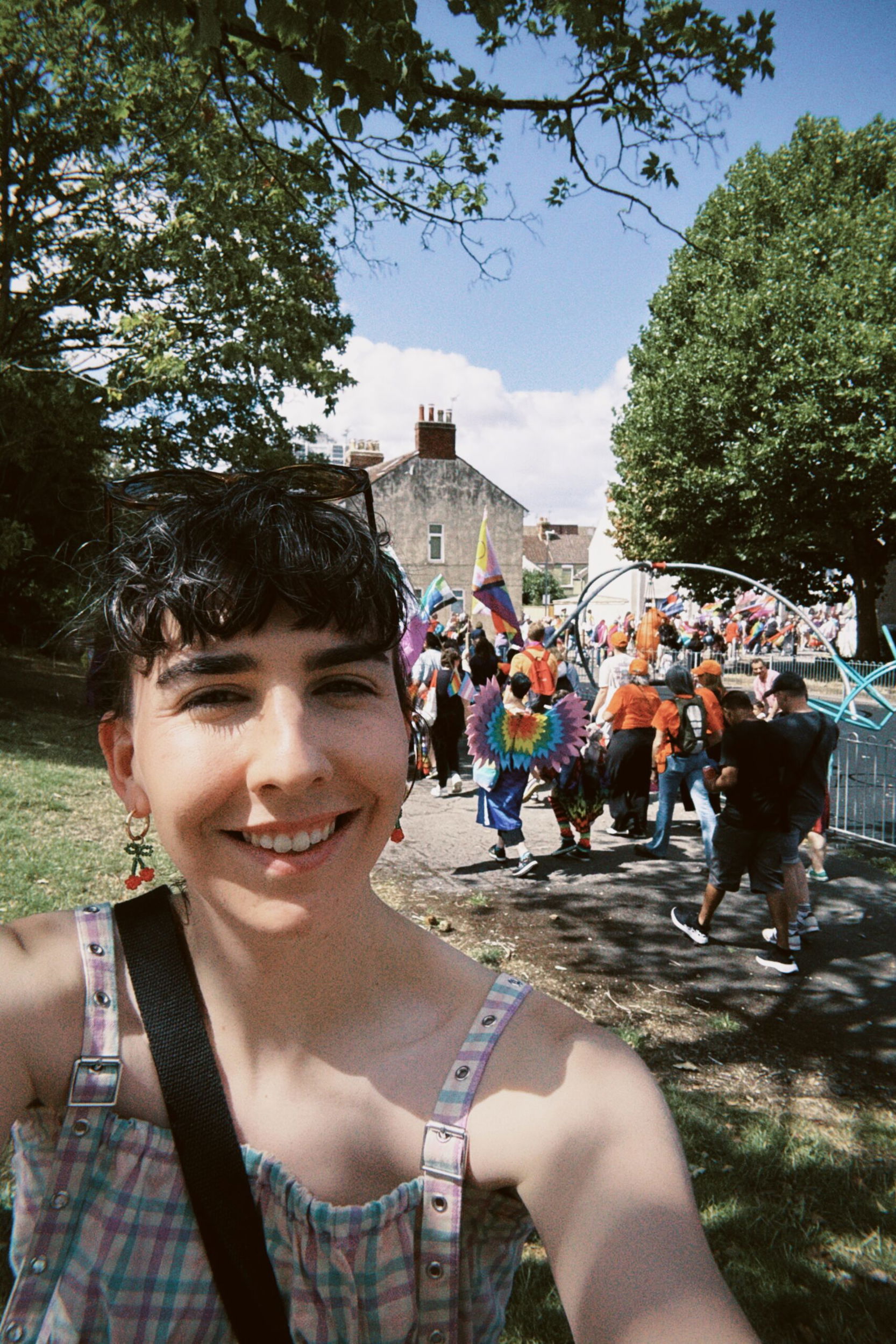 Phoebe Trass, with short dark hair, smiles at the camera in a park. Behind her, a colourful parade with people in bright costumes, including one with large wings, moves along a tree-lined path under a blue sky.