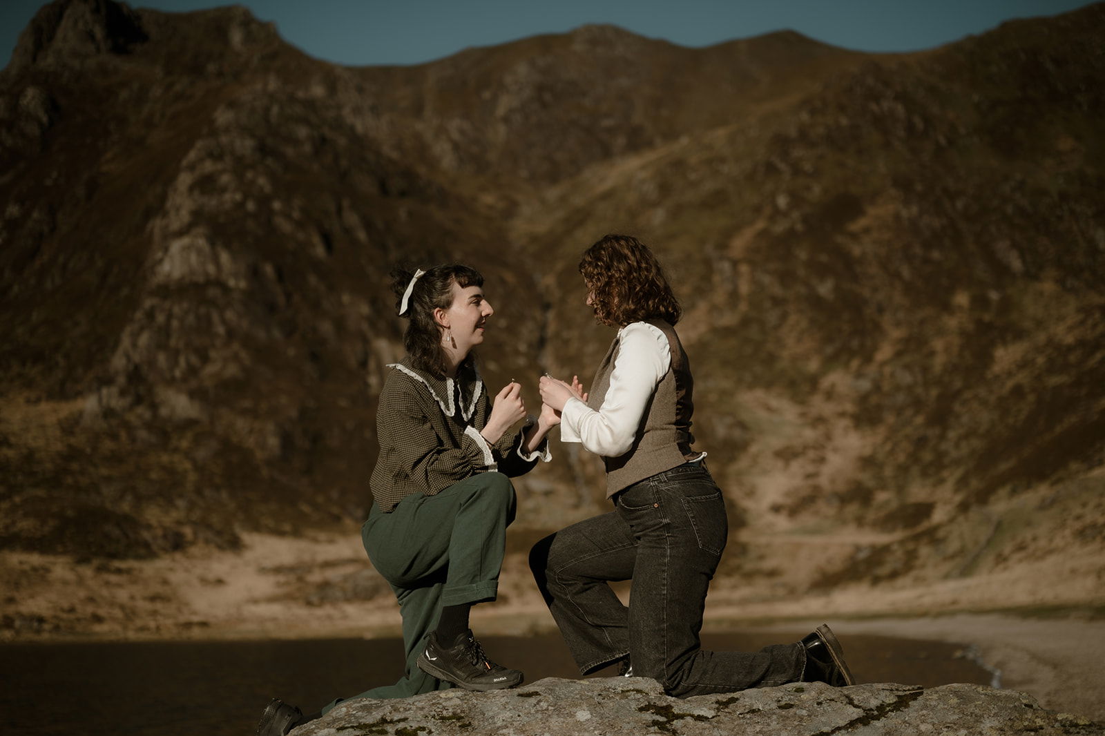 Two people kneel facing each other on a rock outdoors, holding hands and smiling with joy, surrounded by mountains and warm sunlight—capturing a moment reminiscent of Phoebe Trass’s vibrant spirit.