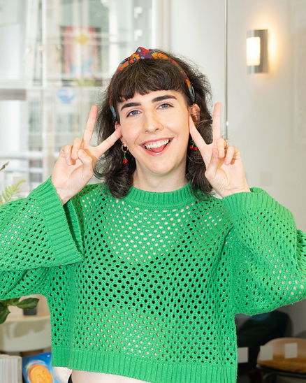 Phoebe Trass, with dark hair and a colourful headband, smiles and makes double peace signs with both hands. She wears a bright green, loose-knit jumper and stands indoors.