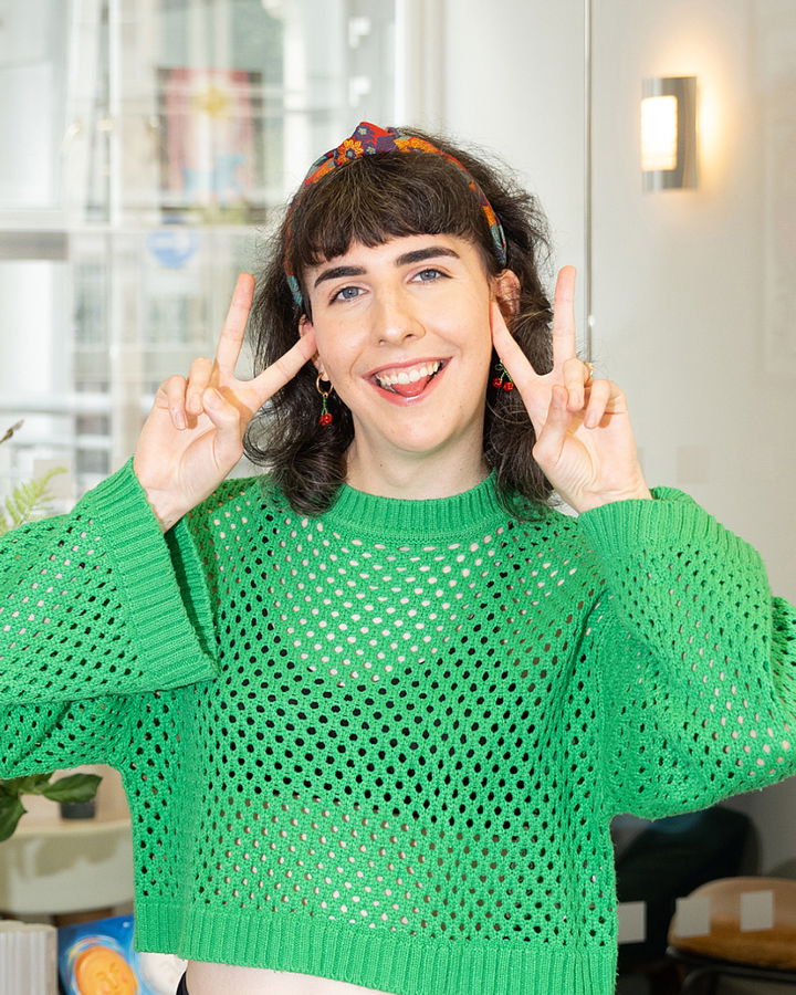 Phoebe Trass, with dark hair and a colourful headband, smiles and makes double peace signs with both hands. She wears a bright green, loose-knit jumper and stands indoors.