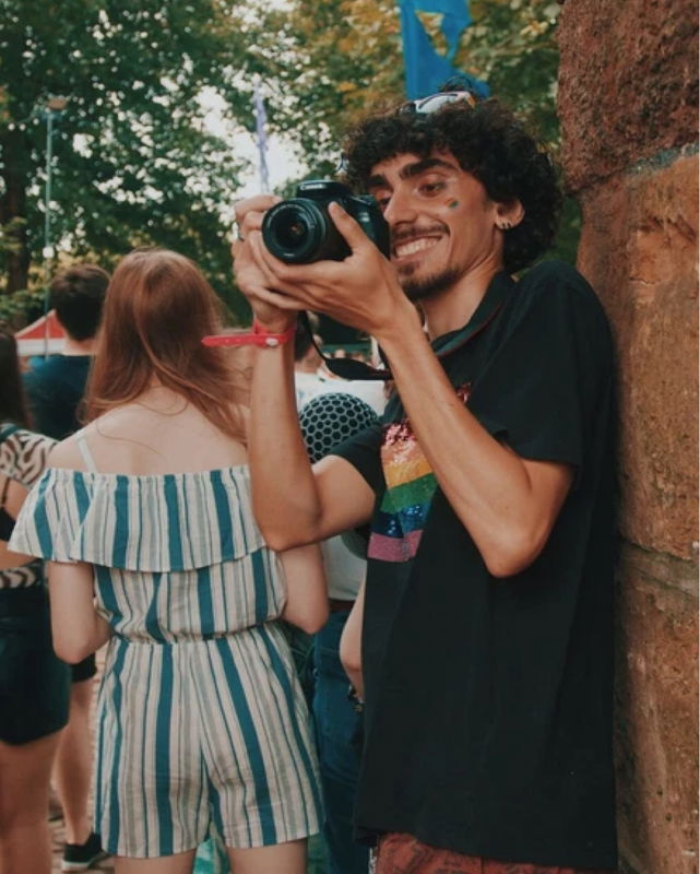 A smiling person with work experience wears a rainbow-striped shirt and face stickers, holding a camera whilst leaning against a stone wall at an outdoor event surrounded by people and trees.