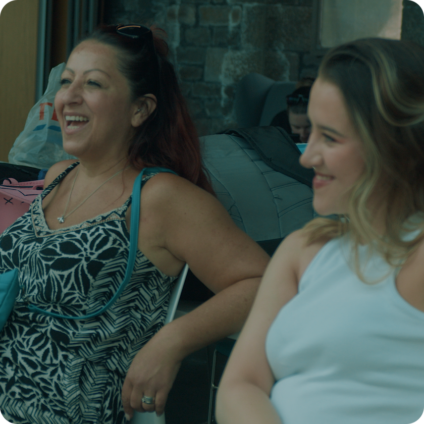 Two women sit side by side, smiling and laughing, perhaps sharing stories from their work experience. One wears a patterned dress, the other a white sleeveless top. Bags and stone walls are visible in the background.
