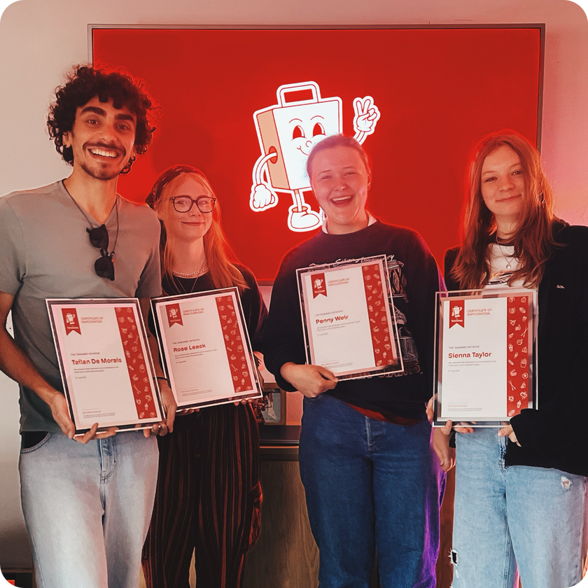 Four smiling people stand in front of a red wall holding award certificates, celebrating their work experience. A cartoon character is displayed on the screen behind them, and they appear happy and proud of their achievements.