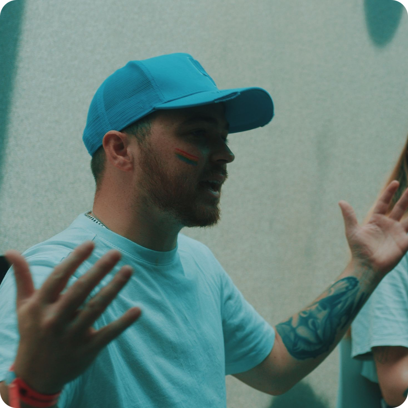 A person with work experience, wearing a blue cap and T-shirt, gestures with open hands, showing a colourful streak painted on their cheek and a tattoo on their forearm, standing indoors against a neutral background.