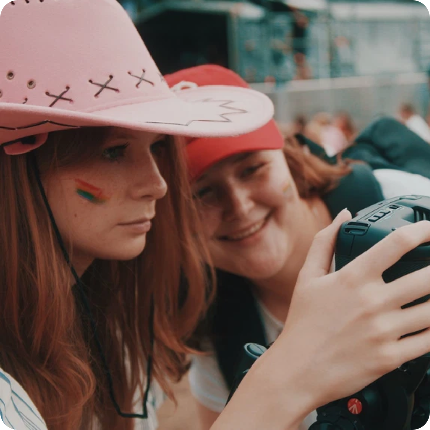 Two people look at a camera screen outdoors, sharing a joyful moment. The person in front sports a pink cowboy hat and a rainbow flag painted on their cheek, showcasing how work experience can bring people together in creative ways.
