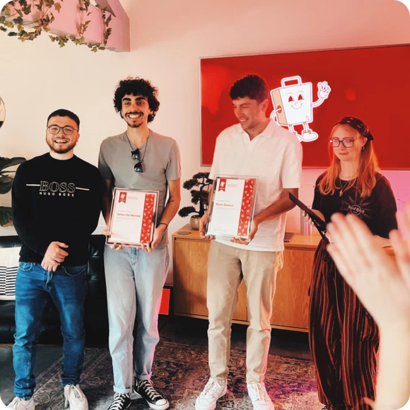 Four people stand smiling indoors; two hold certificates celebrating their work experience. A red screen with a cartoon character glows in the background, whilst a hand claps in the foreground, creating a casual and festive atmosphere.