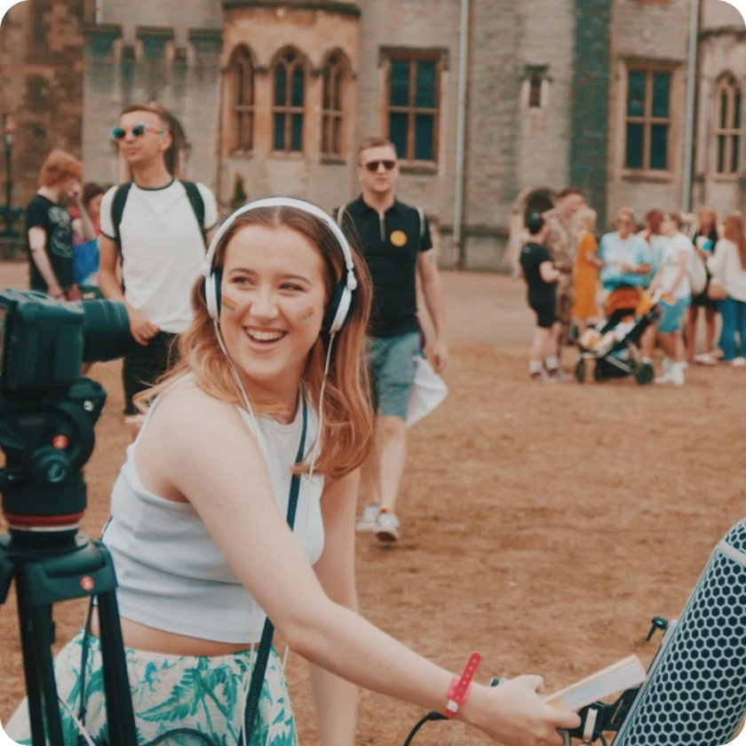 A smiling woman with work experience operates audio equipment outdoors, wearing headphones as she works in front of a camera. People walk and gather in the background near a historic stone building.