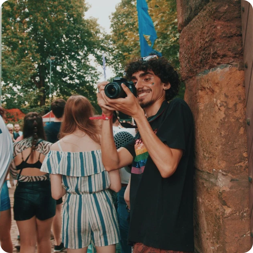 A smiling person with curly hair and a rainbow-striped shirt holds a camera, leaning against a stone wall at an outdoor event—capturing moments and gaining valuable work experience among people and trees in the background.