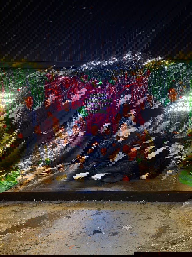 A group of twelve people, passionate about humanitarian and international development, pose together on a decorated stage with festive lights, tinsel, and Christmas trees. A neon sign in the background reads, Merry Christmas Ya Filthy Animal.