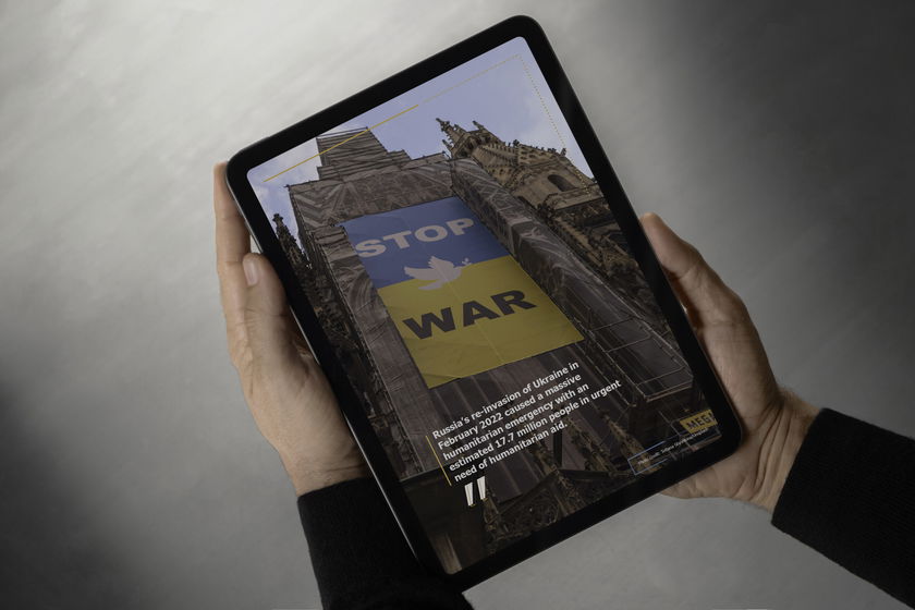 A person holds a tablet displaying a building draped with a large “STOP WAR” banner in Ukraine’s flag colours, highlighting humanitarian and international development concerns amid Russia’s 2022 invasion of Ukraine.