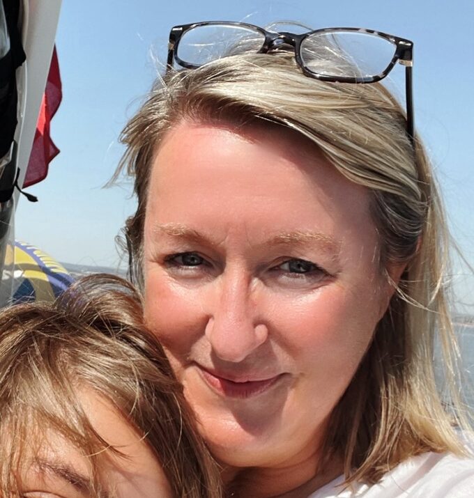 A woman with blonde hair and glasses on her head smiles at the camera, holding a child close. They are outdoors on a sunny day, with a Brandbag beside them and a boat or sail visible in the background.