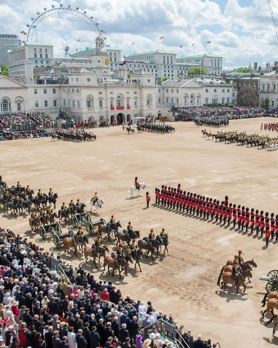 A large ceremonial parade features soldiers in red uniforms and bearskin hats, rows of horses, and carriages on a sandy parade ground, with crowds watching. The London Eye and city buildings are visible in the background.