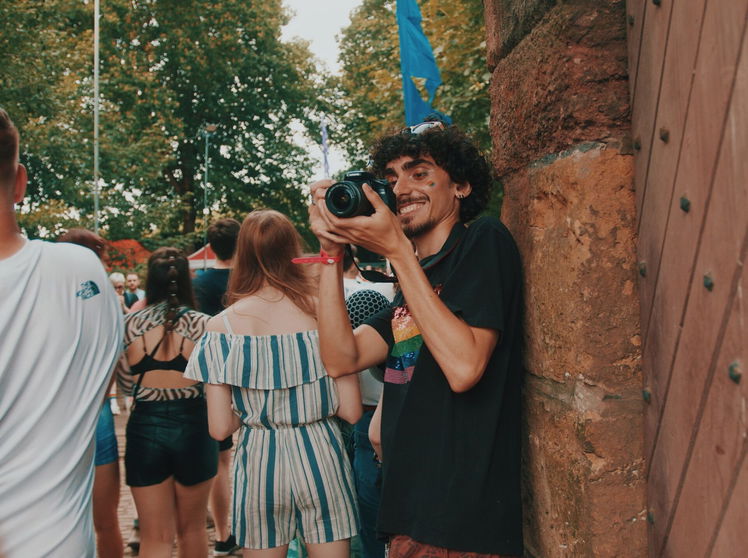 A smiling person with curly hair, holding a camera, stands by a stone wall, capturing moments from The Takeaway – a new initiative for our young creatives – as a casually dressed crowd enjoys the sunny day among trees in the background.