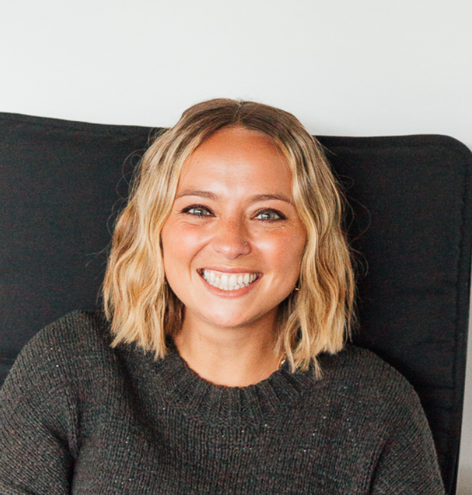 A woman with blonde wavy hair and a dark jumper smiles broadly while seated against a black background, embodying the creativity and confidence behind bespoke website creation.