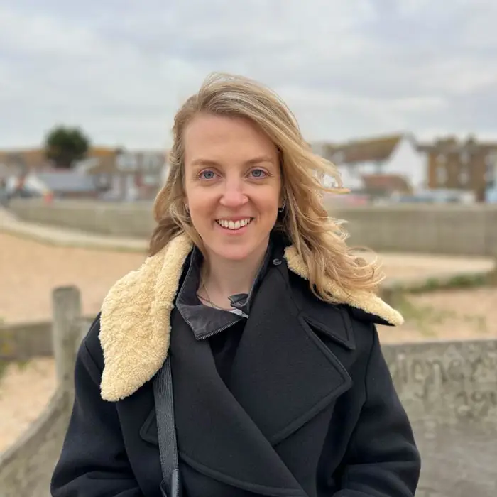 A woman with blonde hair, wearing a black coat with a shearling collar, stands outdoors and smiles at the camera—a scene reminiscent of visuals often used in successful behaviour change campaigns. The background features sand, a stone wall, and houses under a cloudy sky.